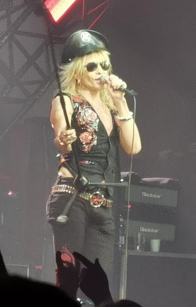 Michael Monroe singing onstage - pvc cap, long blond hair, shades, black waistcoat and black jeans - holding up a mic stand. Basically looking cool as fuck despite being 60, because he is cool as fuck.