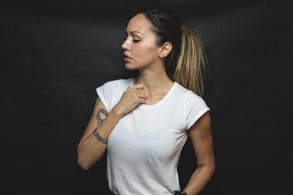 Deborah De Luca is posed with eyes downcast to her right. She sports a white t shirt and a mid pony tail. Her right hand softly grips the top of her shirt collar.