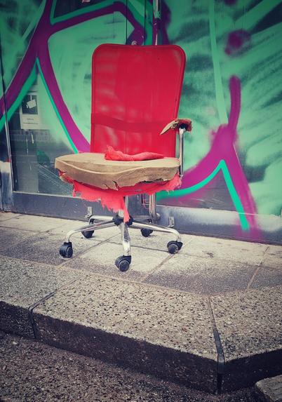 Ein sehr kaputter roter Bürostuhl steht auf einem Treppenabsatz vor einem derzeit geschlossenen Geschäft, dessen Fensterfront mit grünem Graffiti fast vollständig zugesprüht ist.