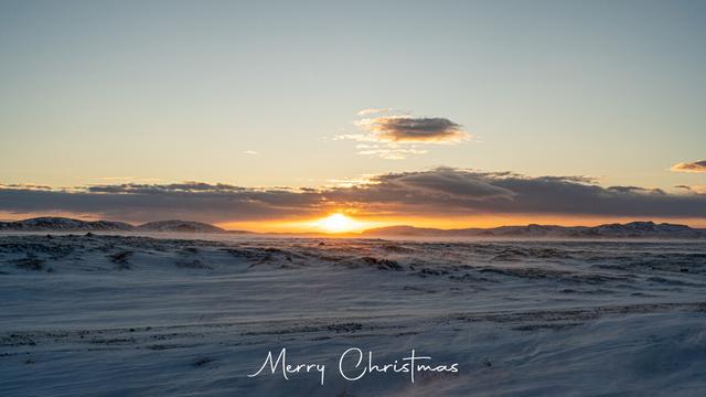 An image from Iceland of a snow covered plain with the setting sun behind it. Stones and lava rocks are poking up through the snow in various areas giving the image great contrast from the white smoothness of the drifting and blowing snow.