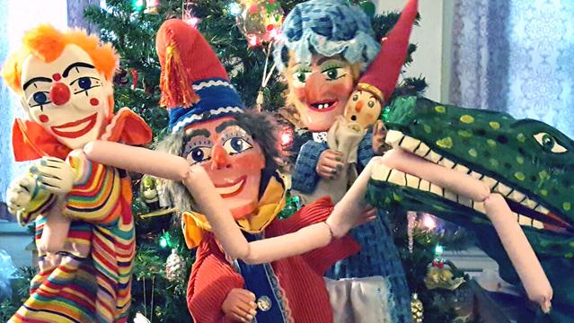 Carved wooden puppets: Punch and Judy, the baby, Joey the Clown, and the crocodile with sausages, in front of a decorated Christmas tree.