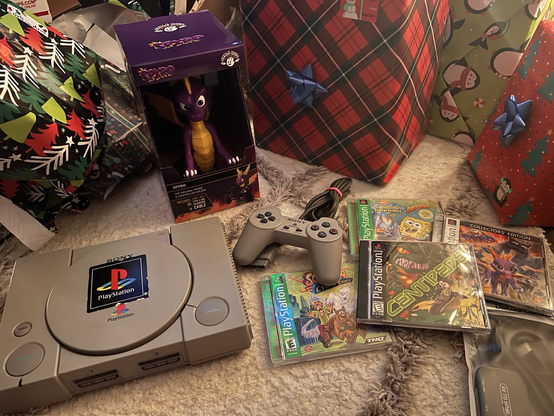 Grey PlayStation 1 console with controller, a Spyro the Dragon (purple dragon) controller holder, Spyro the Dragon, Centipede, Scooby Doo, and SpongeBob game for the PlayStation 1. AV to HDMI cable displayed on white carpet.