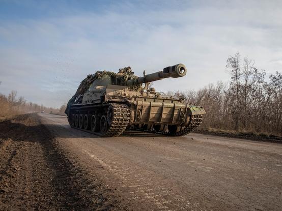 A tank pictured proceeding down tarmaced road marked by other tracked vehicles. Either side of the road are trees with no leavez denoting winter