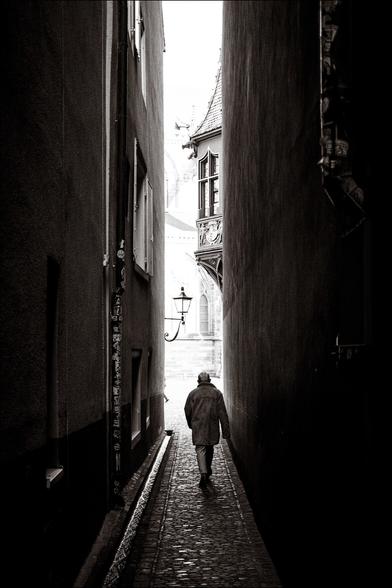 Schwarz-Weiss Bild. Eine enge Gasse in Freiburg an deren Ende ein Mann mit einem Ledermantel läuft. Man sieht die kleinen Steine am Boden schimmern. Ich habe dieses Bild Ende letzten Monats gemacht.