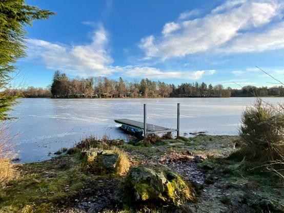 An icy and partly frozen Loch of Aboyne. A small jetty in the foreground. The caravan site can be seen on the far side of the loch.