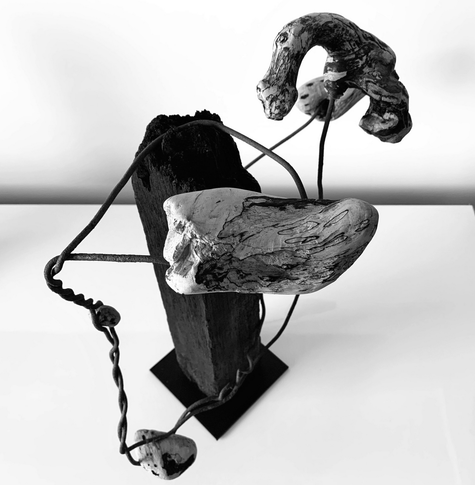 black and white photograph of an abstract sculpture made of driftwood and rusty iron wire.  some pieces of natural colored driftwood are stuck in an irregular helix of wire.  the wire is wound around a central block-shaped black piece of wood, so that the small pieces of wood are trapped like planets in a twisted orbit.  the block of wood stands on a small square plate made of black metal, view from above