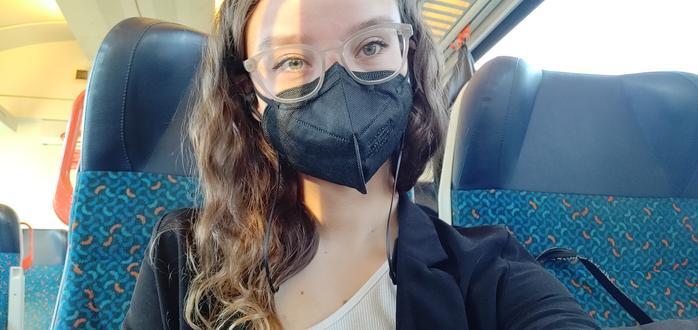 Anne sitzt im Zug und trägt eine schwarze Maske.