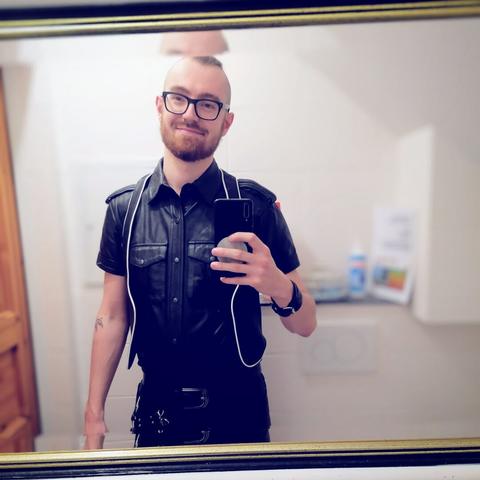 A young man in a bathroom wearing leather pants, shirt and bar vest taking a mirror selfie.