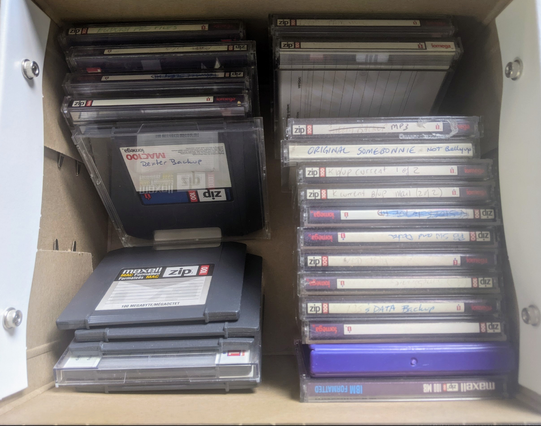 A box of a couple dozen old Iomega Zip disks, most in cases, with barely legible labels describing in vague terms what's backed up on each disk.