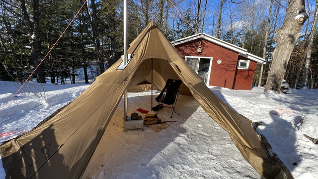 A large tan tipi style tent. Large door is open showing a small wood stove inside with a camp chair in front of it.