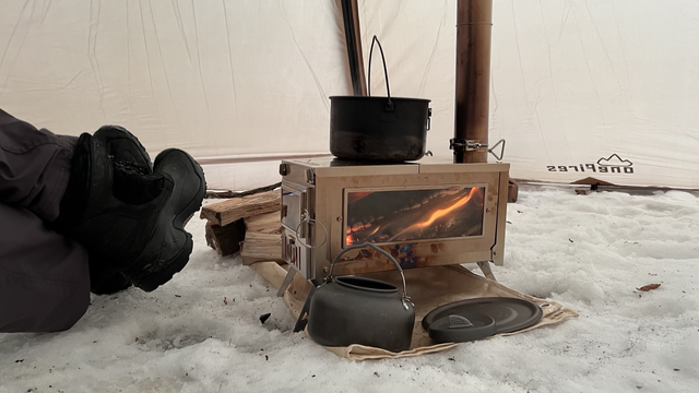 A small metal tent wood stove with a man’s booted feet to the side. A pot is in the stove and the first can be seen through a glass window in the stove.