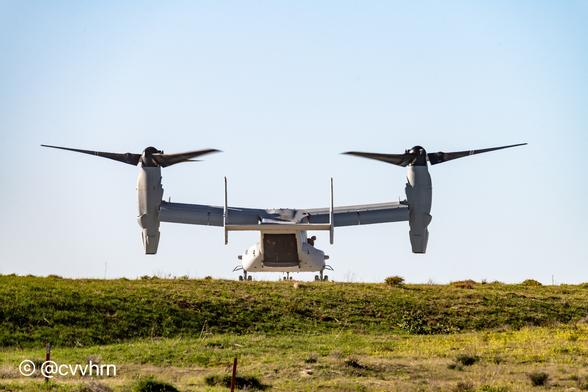 A tilt rotor aircraft is touching down on a grassy knoll. The aircraft has two huge engines, one on each wingtip that can pivot up for helicopter mode, then can transition forward to a fixed wing configuration. you can just see crew and passengers inside as the rear cargo door is open