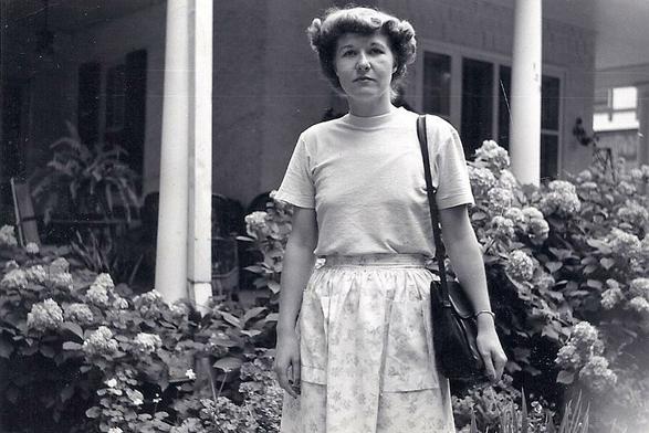 Black and white photo of Jean Bartik, wearing a short sleeved mock turle, and a long skirt. She is facing the camera and is standing in front of a house with flowers all around her.