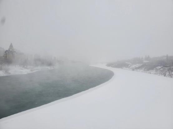 The scene isnshot off a bridge overlooking the South Saskatchewan river. Half the river is iced over. The air is foggy, with the ghost of the Bessborough hotel looming on the left bank , with the right bank only showing winter vegetation.