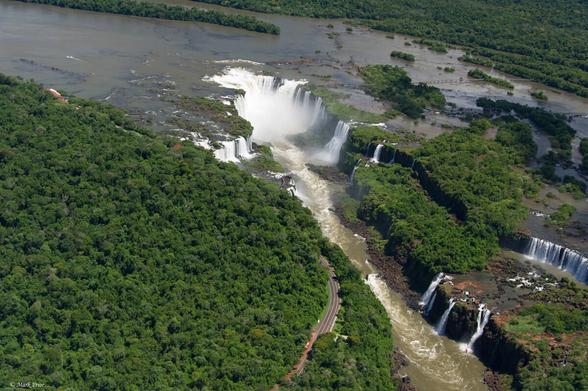 Flying over the Iguazu Falls in a helicopter. The Iguazu River is spread wide across the top of the frame and then it hits the Devil's Throat, a horseshoe shaped waterfall creating a cauldron of white water and spray. To the left of it are more waterfalls on the Brazilian side of the river which are spectacular when up close but look tiny when compared to the Devil's Throat from above. Stretching away from the Devil's Throat to the lower right hand corner are more waterfalls in two levels along the Argentine border. In wet season I suspect it is a curtain of water but in December there are separate waterfalls. On the left of the frame is thick green jungle but a small road cuts a narrow path between the river and the jungle. The terracotta coloured roofs of some buildings are seen poking out of the jungle canopy. To the right of the falls the river continues and feeds further, out of shot, waterfalls. There is a boardwalk on the Argentine side skipping across some small islands in the river until it reaches an observation deck overlooking the Devil's Throat.