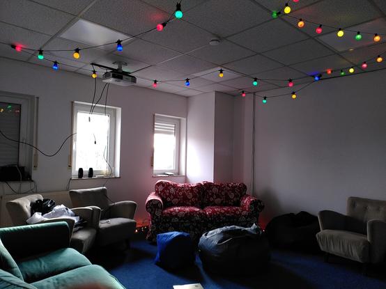 Raum mit Couchen, Sesseln uns Sitzsäcken. Auf dem Boden ein blauer Teppich. An der Decke hängen bunte Lichterketten.
