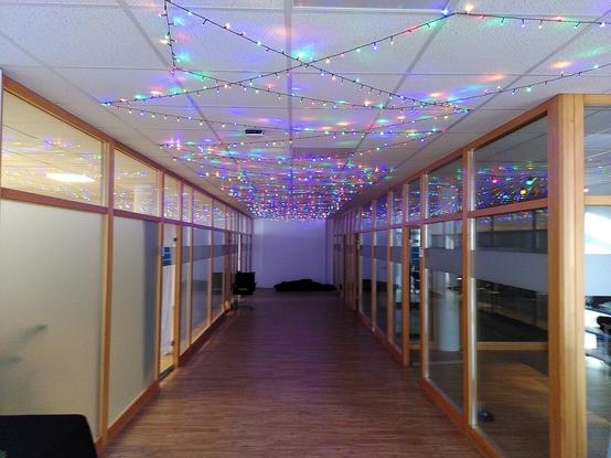 Blick in einen Flur. Links und rechts sind Glaswändende. An der Decke sind im Zickzack Lichterketten mit winzigen, bunten LEDs gespannt.