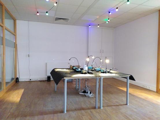 Blick in einen Raum. In der Mitte steht ein quadratischer Tisch mit Lötstationen und Schwanenhals Lampen. An der Decke eine bunte Lichterkette.