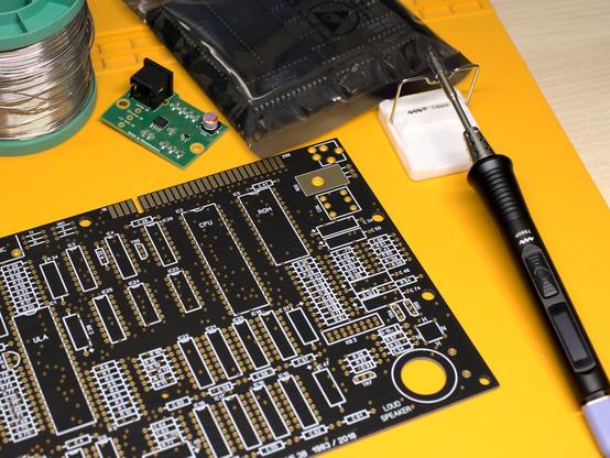 An empty ZX Spectrum Issue 3 replica board, black solder mask and ENIG finish. There is also a soldering iron and a spool of solder, a S-Video module, and an ESD safe bag with chips.