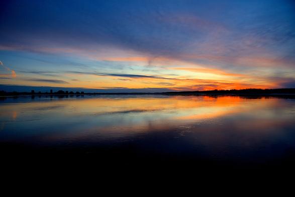 The fading sunlight from just after sunset on the Ottawa River casts a fiery reddish-orange on the right side of the photo and stretches out to blue as light crosses to the left side of the image. Cloud cover adds to the drama and the scene is mirrored on the reflective surface of the river.

The water at the bottom of the image is almost black.