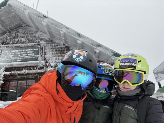 My twin boys and I standing in front of cabin that says Little Whiteface Mountain in gray wintery conditions in our ski helmets, goggles, and jackets