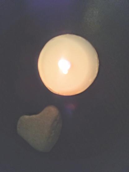 Colour photograph in low lighting. Pictured is a white tealight candle, lit, beside a stone that is naturally heart-shaped.