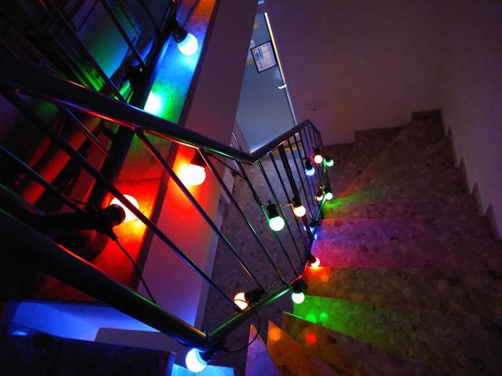 Blick von oben, ein Treppenhaus herunter. Um das Geländer ist im dunklen eine bunte Lichterkette gewickelt.