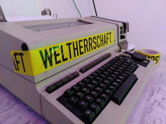 Ein Fernschreiber T1000 von Siemens auf einem Tisch. Darauf ein gelbes Klebeband mit schwarzer Aufschrift "Weltherrschaft"