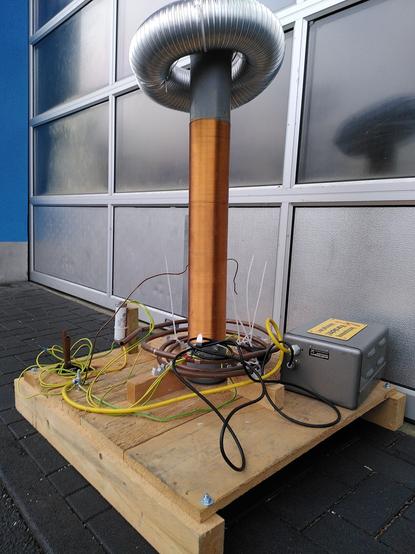 Bild auf Kniehöhe. Man sieht eine Teslaspule, montiert auf eine Holzpalette. Fein gewickelter Kupferdraht auf einem grauen Abflussrohr, darüber ein metallbeschichteter Torus. Am Boden eine aus Kupferrohr gebogene Spule und ein Hochspannungstransformator