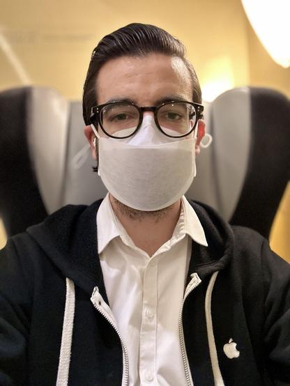 Selfie, portrait mode; I am sitting on a train in a comfy seat; I’m wearing a white Apple Mask, a white shirt and a black hoodie with an embroidered Apple logo on the left