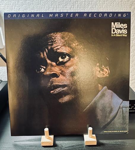 Photo of a record album cover, black background dominated by the face of Miles Davis, eyes wide, looking up and to the left. 

In the top right hand corner in white lettering, Miles Davis. Beneath this, in yellow, In A Silent Way. The banner across the top of the album cover reads, ‘Original Master Recording’.