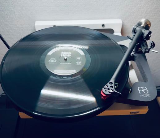 Photo of a record spinning on the skeletal plinth of a Rega P8 turntable. The centre label reads, ‘Miles Davis’. The other lettering is blurred and illegible. 

The tone arm, red cartridge, traces the first track.