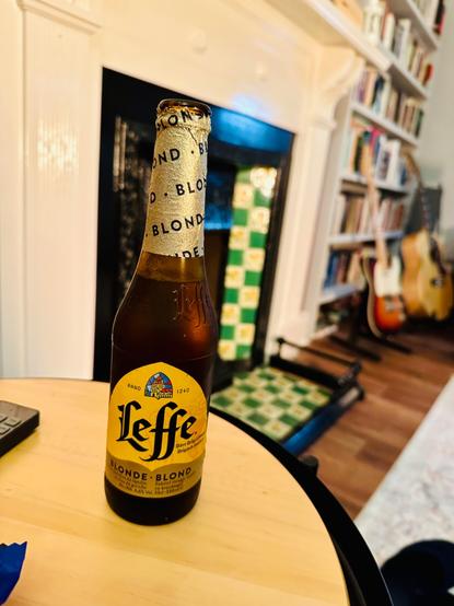 Photo of a bottle of Leffe blonde beer in a pine coloured table. 

In the background, there is a fire place tiled yellow and green, paint work in jet black. 

To the far right of the photo, just out of focus, two guitars, a sunburst Fender Telecaster, and  acoustic Gibson superjumbo, sit propped against a bookshelf overladen with books.