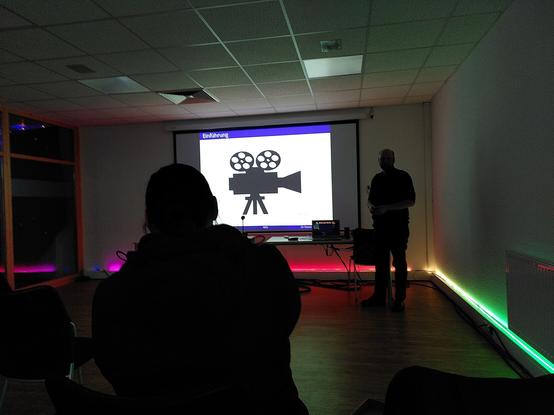 Dunkler bunter Raum, Blick auf eine Leinwand, im Vordergrund eine Silhouette einer Person. Der Vortragende ist auch wie ein Scherenschnitt zu sehen. Das Beamerbild zeigt ein Filmkamera-Icon auf weißem Hintergrund.