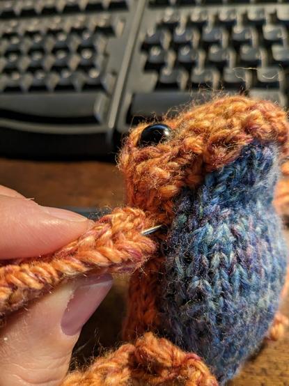 orange knit frog with a blue belly and a darning needle buried in its armpit