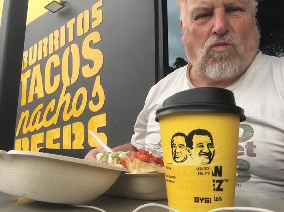 A person at a table outside a Mexican restaurantwith Bargasse (clamshell) takeaway container with food and a takeaway coffee cup. Person does not look very happy.