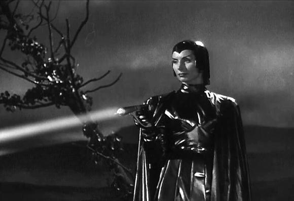 Black and white picture from the movie "Devil Girl From Mars" showing a lady, all in black including a black helmet covering her hair. She is firing a ray gun at something off screen and the beam can be seen. The background is a dark landscape of black ground, a spare tree is tilted sideways with black leaves and bare branches.