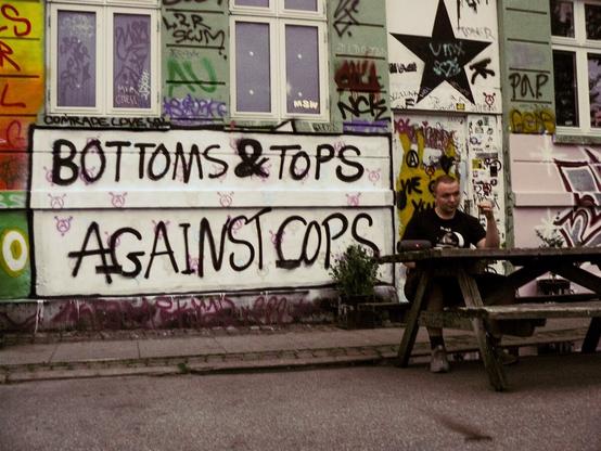 "Bottoms & Tops Against Cops" written on a graffiti covered wall and a person is sitting on a bench