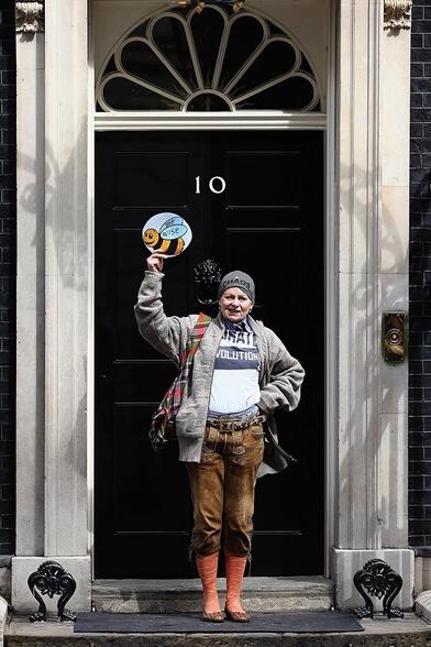 Vivienne Westwood in a gray hat that says "CHAOS", holding up a sign that says "bee wise" in front of the black door of 10 Downing Street in London.