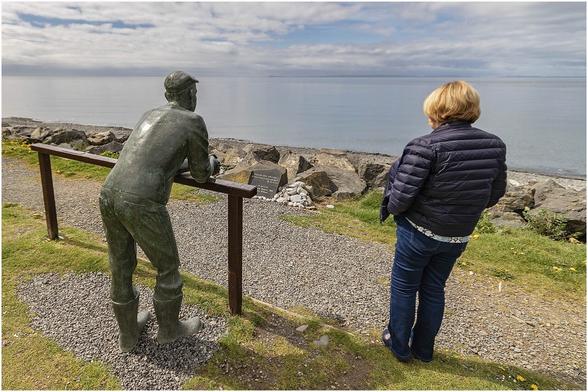 Statue of man leaning on a wooden beam looking out to sea. There is a real woman standing nearby. Is she looking at the statue or staring into the distance..