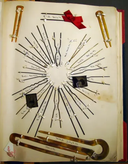 Page in a scrapbook: at the center, hairpins have been sewn onto it so as to form a heart shape, and  names (of their owners?) are written inside each pin.  There are larger pins at the top and bottom of the page, the one at the top having a red ribbon tied in a box affixed to it.