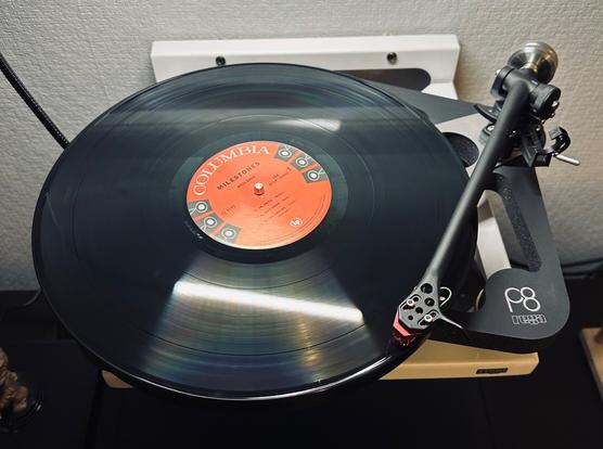 Photo of a record spinning on the skeletal plinth of a Rega P8 turntable. 

The centre label reads, ‘Columbia, Milestones’ above the spindle. To the left and the right of the spindle are three prints of the Columbia logo (this is a replica of the ‘Six-eye label’ - so named after the six prints of the Columbia logo). 

The tone arm, with a hint of a red cartridge, is tracing the first track. 

The turntable is resting on a white wall bracket.