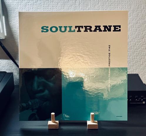 Photo of a record album cover. The cover is divided into three quadrants. 

The top half of the cover is white with the title of the album printed in blue and black type: ‘Soultrane’. 

The lefthand corner has a photo of the lower part of John Coltrane’s face as he blows into the mouthpiece of a saxophone. 

The bottom right hand corner is coloured pale blue. Small lettering runs vertically through the right hand corner and into the upper half of the cover, reading, John Coltrane Prestige 7142.