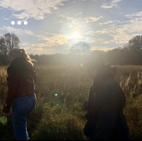 Auf dem Foto sind zwei Personen zu im Gegenlicht abgebildet, die nach links über ein Feld gehen. Im Hintergrund steht die Sonne tief hinter einem Waldrand. Beide Personen haben winterliche Kleidung an und leuchten im herbstlich-winterlichen kühlen Abendlicht.