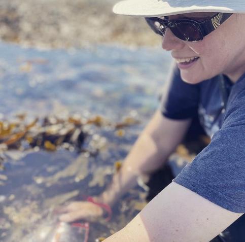 Elizabeth sits in a rockpool with her hands in the water. She is wearing a beige hat and sunglasses. She has a big smile and is clearly very happy to be there.