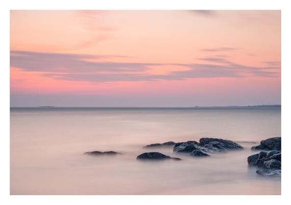 A still sea with rocks in the foreground with a colourful, yet delicate, sunset.