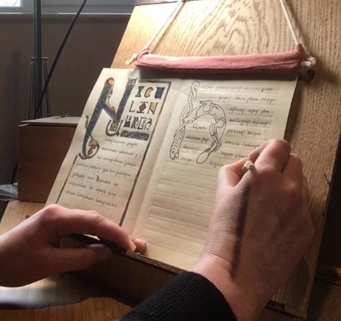 A sheet of parchment on a writing stand. It is folded into two pages. Someone is writing with a quill on the right-hand page. The left page is finished and shows an illuminated poem.