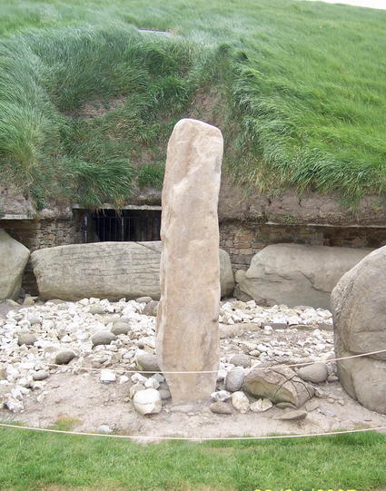 Tall limestone pillar in foreground surrounded by rounded rocks. Long oblong kerbstones in the background marking the base of a mound covered in grass.