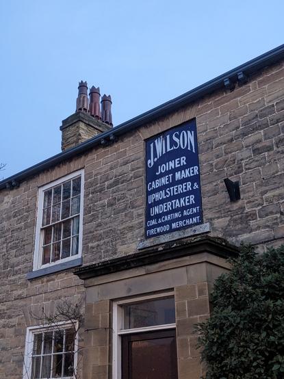 Painted sign on the front of an old stone building. "J Wilson - Joiner - Cabinet Maker - Upholsterer & Undertaker - Coal & Carting Agent - Firewood Merchant"