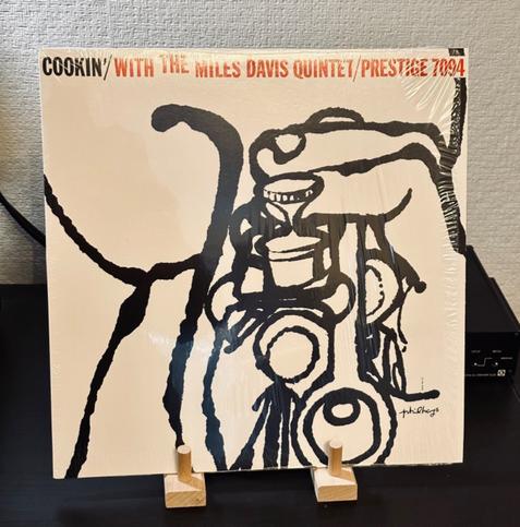 Photo of a record album cover. The cover is white with an ink sketch of the valves of a trumpet, fingers pressing the keys. 

The typeface across the top of the sleeve reads, Cookin’ with the Miles Davis Quintet/Prestige 7094.